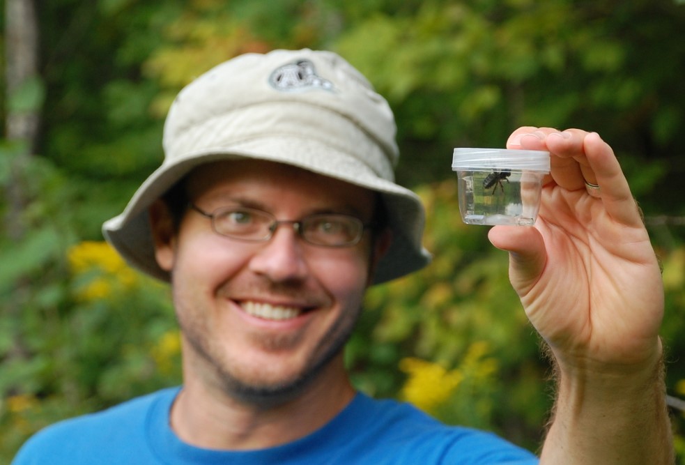 Photo of Jay holding a bumble bee in a specimen jar outdoors.